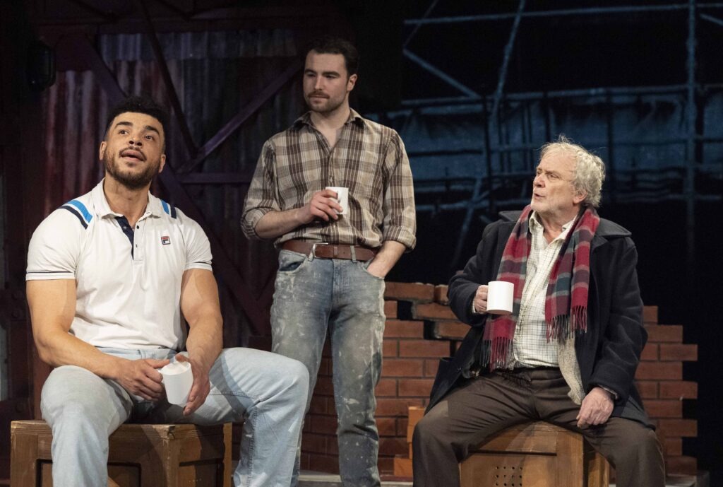 Jurell Carter (Loggo), George Caple (Chrissie) and Ged McKenna (George). George and Loggo sat on wood boxes. Chrissie stood up with cup of tea.