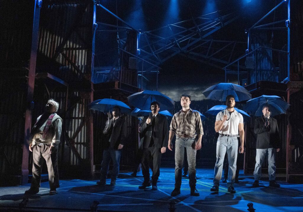 Reiss Barber as Snowy Mallone (Centre) and The 2025 Tour Cast of Boys from the Blackstuff - all stood on stage most holding black umbrellas.