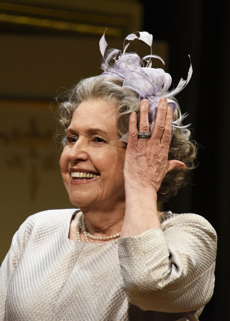 ANNE REID as The Queen, wearing white fascinator