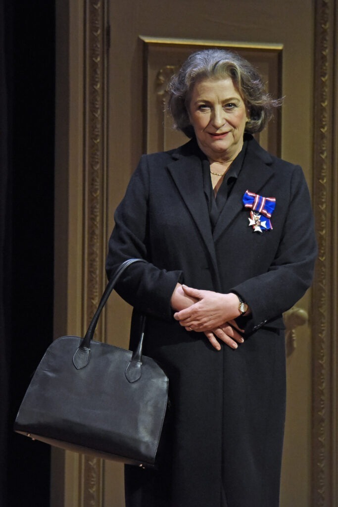 CAROLINE QUENTIN as The Dresser, wearing all black carrying black handbag
