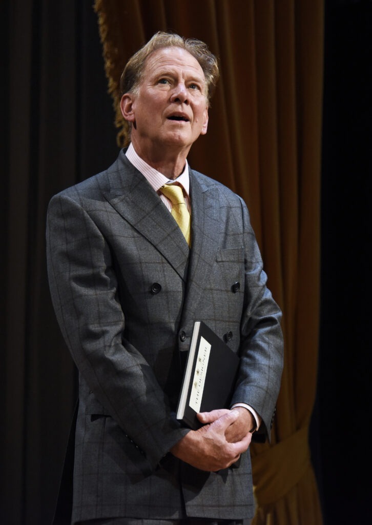 JAMES WILBY as The Designer, wearing grey suit with yellow tie. Carrying sketch book.