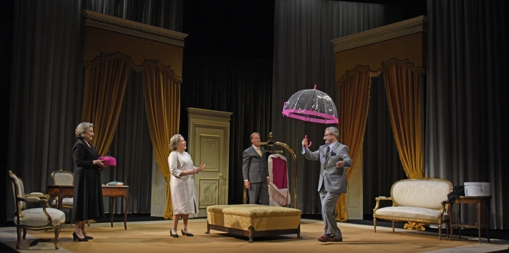 The Dresser, The Queen, The Designer, The Milliner. All looking at The Milliner carrying an open clear plastic umberella with pink rim.