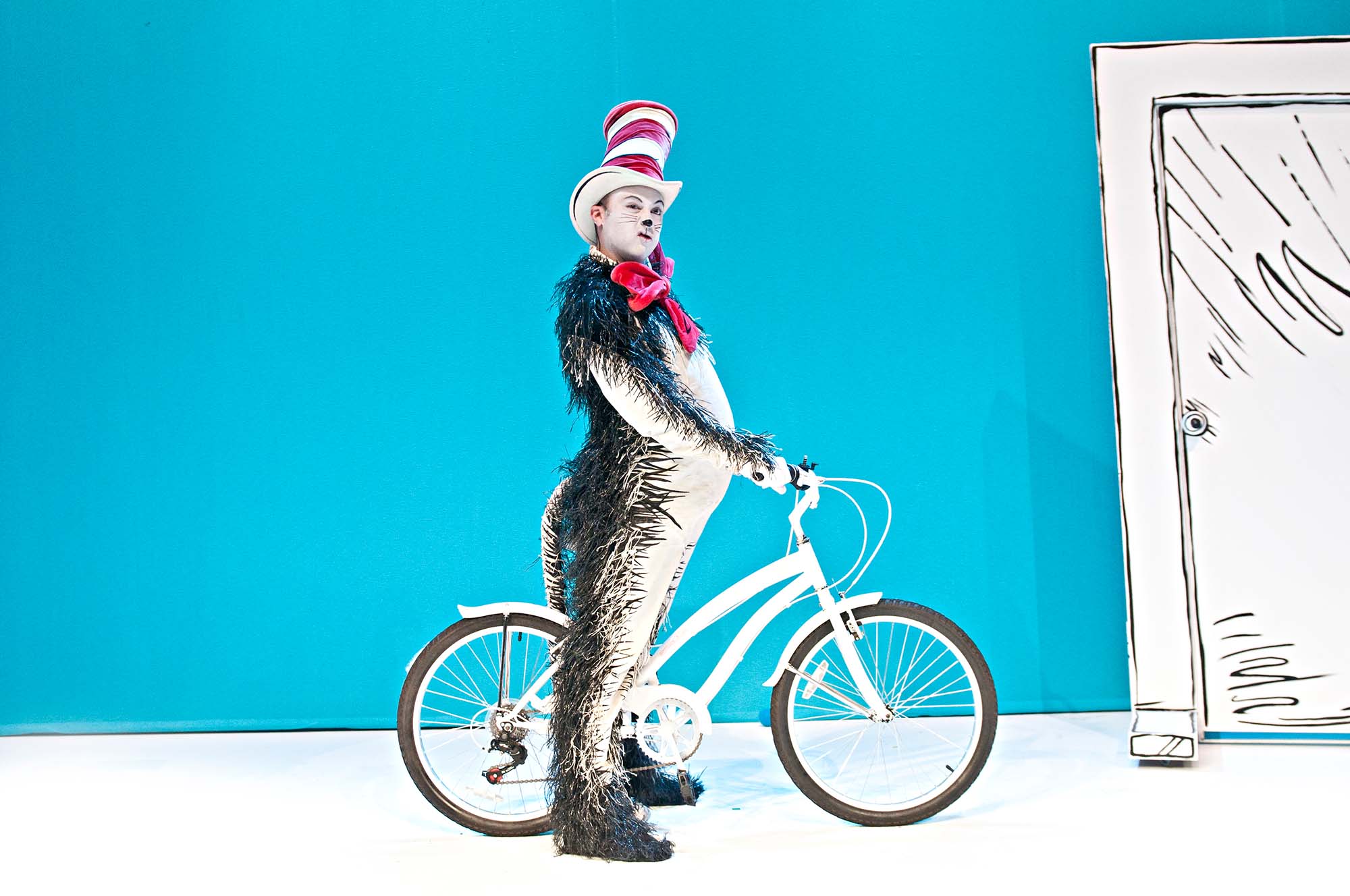 The cat in the hat, in costume, riding a bicycle.